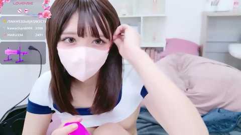 A video of a young Asian woman with straight brown hair, wearing a white surgical mask and blue top, lying on a bed in a modern bedroom, holding a pink vibrator.