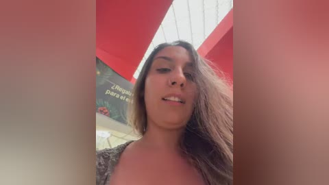 Video of a young woman with long brown hair, light brown skin, and a casual outfit, standing under a red and white structure with text in the background.