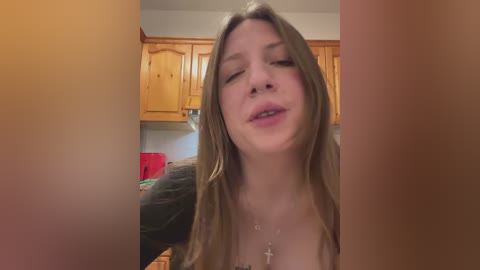 Video of a young Caucasian woman with long, light brown hair, wearing a black top and a cross necklace, in a domestic kitchen setting with wooden cabinets.