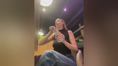 Video of a light-skinned woman with long brown hair, wearing a black sleeveless top and jeans, drinking water from a bottle, seated indoors with a warm, orange wall and modern lighting.