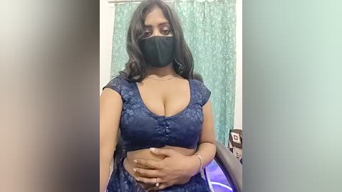Video of a South Asian woman with medium-dark skin, wearing a black face mask, navy blue floral crop top, and black pants, standing in a small, dimly lit bathroom with a teal shower curtain.