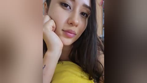 Media: Video of a young woman with long dark hair, light skin, and a small tattoo on her wrist, wearing a yellow top, resting her chin on her hand, looking introspective.