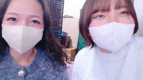 Video of two Asian women with short brown hair, wearing white surgical masks, blue sweaters, and green backgrounds; one is smiling, the other neutral.