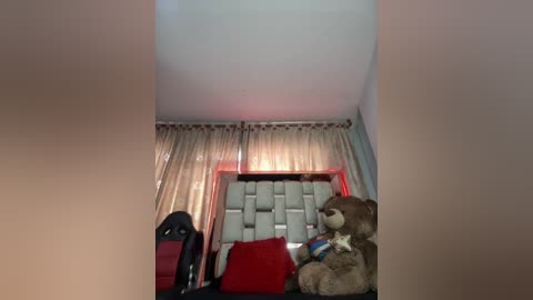 A video of a cozy bedroom with a plush teddy bear, red and black pillows, a tufted headboard, and sheer curtains in a muted color palette.