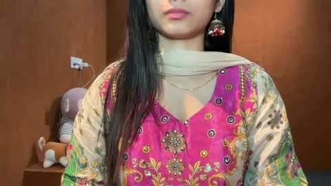 Video of a woman with long black hair, wearing a vibrant pink embroidered shalwar kameez with intricate floral designs, against a warm brown wall, with a stuffed toy in the background.