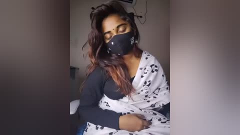 Media: A video of a young South Asian woman with long, wavy dark hair, wearing a black face mask, black long-sleeved top, and a white sari with black floral pattern, sitting indoors with neutral background.