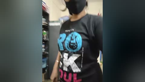 Video of a person in a black \"Rolling Stones\" t-shirt with a blue \"ROCK\" logo, wearing a black face mask, in a grocery store aisle with shelves and products visible.