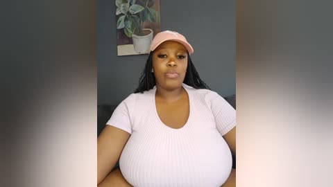 Video of a plus-size Black woman with long black hair, wearing a pink cap and ribbed white top, sitting indoors against a gray wall with a potted plant and framed artwork in the background.