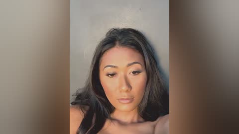 Video of a young Asian woman with long, wavy dark hair, wearing subtle makeup and a soft, neutral expression, set against a blurred, neutral background.