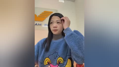 Video of a young Asian woman with long black hair, wearing a blue fuzzy sweatshirt with a cartoon duck face and yellow eyes, looking thoughtful, in a beige room with a DHL logo.