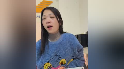 A video of an Asian woman with long black hair, wearing a blue knit sweater with cartoon eyes, smiling and laughing in a modern, brightly lit room.