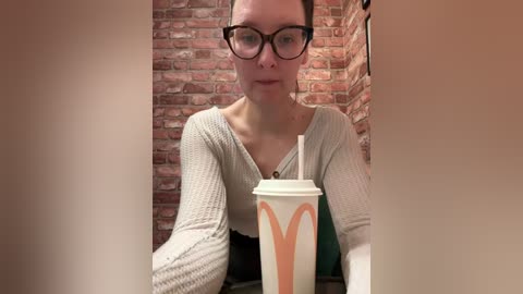 Video of a fair-skinned woman with glasses, wearing a light gray, ribbed sweater, seated in a brick-walled coffee shop, holding a McDonald's cup with a red M logo.
