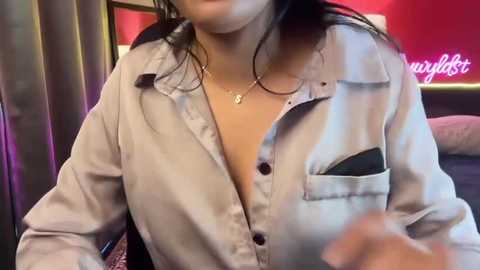 A video of a woman with fair skin, medium build, wearing a partially unbuttoned light blue shirt, revealing a black bra, and a gold necklace. Background shows a red wall with \"Way\" written on it.