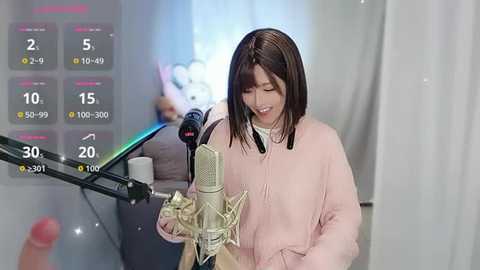 Media: Video of a smiling, East Asian woman with straight, shoulder-length brown hair, wearing a light pink sweater, singing into a green microphone. Background shows digital overlays of a video game, with a blurred, white curtain.