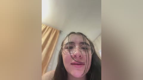 Video of a young woman with long, dark hair, wearing clear-framed glasses, and a neutral expression. The background features beige curtains and a white wall.