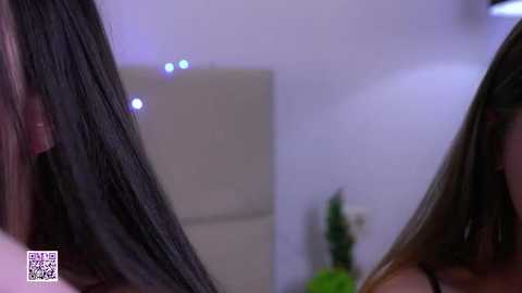Media: Video of two Asian women with long, straight black hair in a modern, dimly-lit room. The blurred background includes a potted plant and a white wall with indistinct lighting.