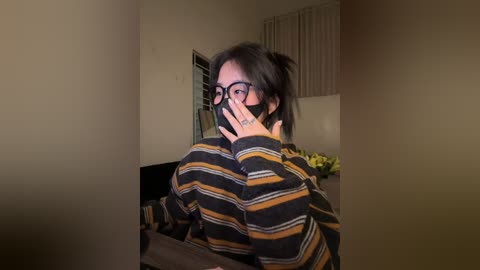 A video of an Asian woman with short black hair, wearing black-framed glasses and a black mask, in a striped sweater, sitting in a dimly-lit room with beige walls and a window with vertical blinds.