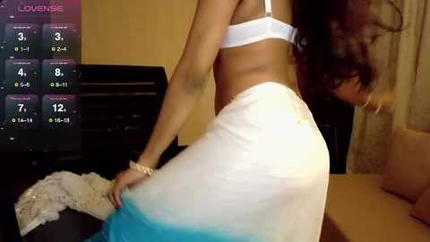 A video of a woman in a white bikini, with long dark hair, adjusting her top in a dimly lit bedroom. The background shows a TV displaying a \"Love Island\" countdown.