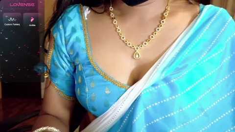A video of a woman with medium skin tone wearing a turquoise saree with gold embroidery, a gold necklace, and a black face mask.