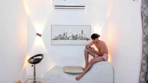 Video of a shirtless, lean, fair-skinned man with short dark hair, wearing pink shorts, sitting on a white couch, looking at a phone, in a modern, minimalist room with white walls and city skyline art.
