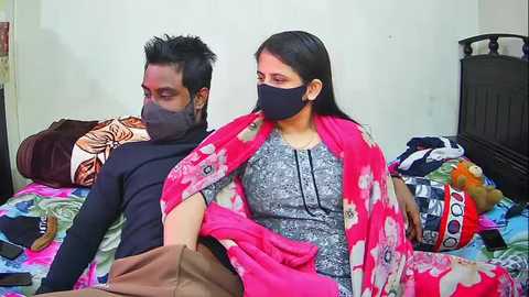 Media: Video of a South Asian couple lying on a colorful bedspread, wearing masks; the man has short black hair and a beard, the woman has long black hair, both dressed in casual attire.