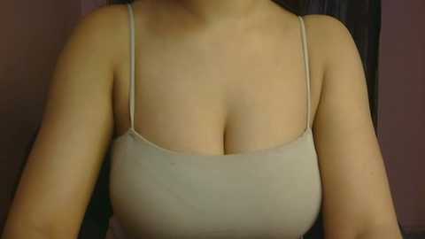 A video of a light-skinned woman with medium-sized breasts wearing a beige, thin-strapped spaghetti-strap top. The background is blurred, featuring a dark-colored wall. The image focuses on her chest area, capturing the fabric's texture and her neckline.