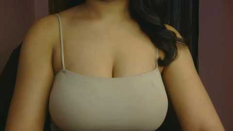 Video of a woman with medium skin tone, wearing a beige spaghetti-strap tank top that highlights her large breasts, with dark hair partially visible in the background.
