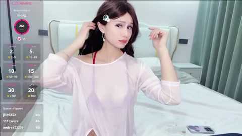 A video of an East Asian woman with fair skin, dark hair, and a sheer white blouse, adjusting her hair in a hospital room with a bed and green curtains.