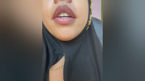 Video of a woman with light brown skin, wearing a black hijab with gold accents, her lips slightly parted, showcasing a glimpse of her teeth.