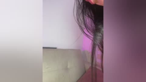 Video of a woman with long, straight black hair, partially obscuring her face, standing in front of a white wall with a purple light casting a glow.