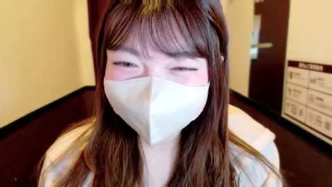 Media: Video of a young Asian woman with straight brown hair, wearing a white face mask and light jacket, indoors against a beige and dark brown wall, with a signboard in the background.