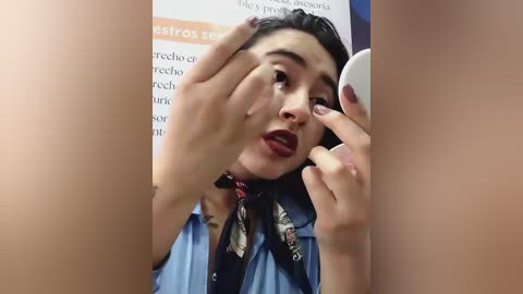 Video of a Latina woman with medium skin tone, dark hair, and full lips. She wears a blue shirt, black scarf, and is on a phone, applying makeup to her left eye. Background includes a poster with Spanish text.