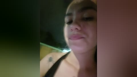 Video of a close-up, slightly blurred woman with light skin and dark hair, wearing a black top, with a tattoo on her shoulder, in a dimly lit room.