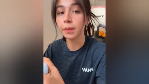 A video of a young woman with light brown skin and straight black hair, wearing a dark blue Vans t-shirt, holding a white remote control. The background is blurred, showing a beige wall and a chair.