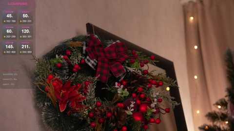 Video of a festive Christmas wreath with red berries, pine branches, and a red plaid bow on a beige wall. Background includes a lit string of fairy lights and a partially visible Christmas tree.