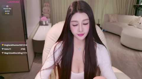 Media: Video of an Asian woman with long black hair, wearing a white top, sitting in a beige chair in a modern living room with a beige sofa and white curtains.