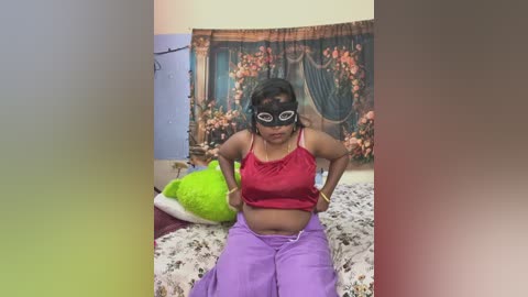 Video of a young woman with dark skin, wearing a red crop top, purple pants, and a black mask, posing on a bed with a floral tapestry and green pillows in a cozy room.