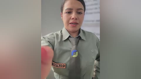 Video of a Latina woman with short dark hair, wearing a gray security uniform with a badge, speaking to a blurred figure in a neutral-toned room.