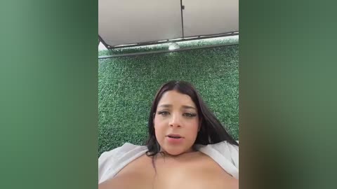 Video of a young woman with long black hair, lying on a green artificial turf, wearing a white top, looking up with a neutral expression.