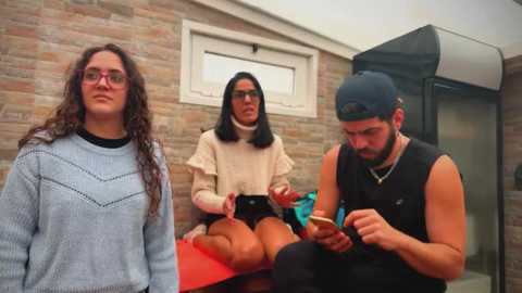 Video of three people: a curly-haired woman in glasses, a man in a black tank top, and a woman in a beige sweater, seated on red and blue chairs against a brick wall.