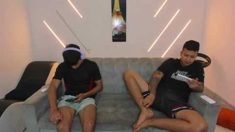 Video of two young men with medium skin tones, wearing headphones and casual clothes, sitting on a gray sofa in a minimalist room with modern lighting and a poster on the wall.
