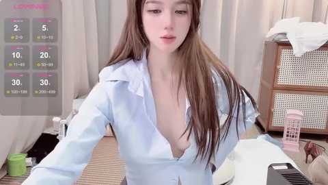 Video of a young Asian woman with long brown hair, wearing a light blue, open-collared blouse, sitting at a cluttered desk in a brightly lit room.