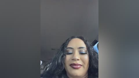 Video of a smiling, light-skinned woman with long, wavy black hair, wearing dark lipstick and subtle eye makeup, against a plain, dark background.