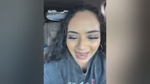 A close-up video of a joyful, light-skinned woman with curly black hair, wearing a grey sweatshirt and hoop earrings, grinning widely in a dimly lit room.