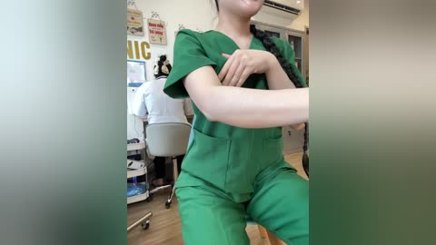 Video of a smiling, light-skinned female nurse in green scrubs with short hair, arms crossed, in a modern hospital room with a desk, computer, and posters in the background.
