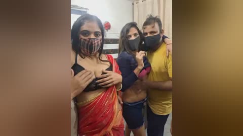 Video of three individuals wearing face masks, with two women in traditional Indian clothing and a man in casual attire, standing indoors.