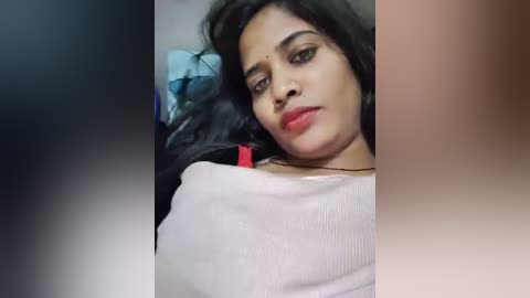 Video of a South Asian woman with medium-dark skin, long black hair, and red lipstick, wearing a white shirt, lying on a bed, with blurred background.