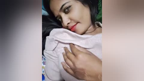 Video of a South Asian woman with medium brown skin, dark hair, and red lipstick, lying on a bed, wearing a light pink shirt, with a man's hand on her breast.