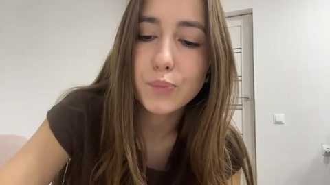 Video of a young woman with long, straight brown hair, making a kissy face. She has a light skin tone and wears a black t-shirt. Background shows a white wall and a partially visible door.