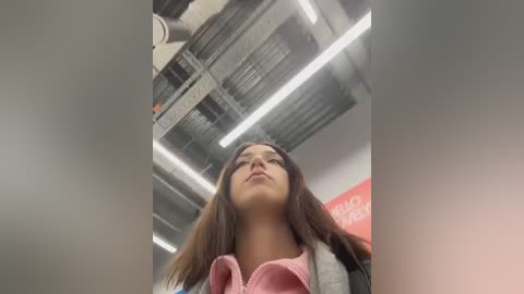 Video of a young woman with long brown hair, wearing a pink hoodie, looking up towards the ceiling with fluorescent lights and exposed pipes in an industrial setting.